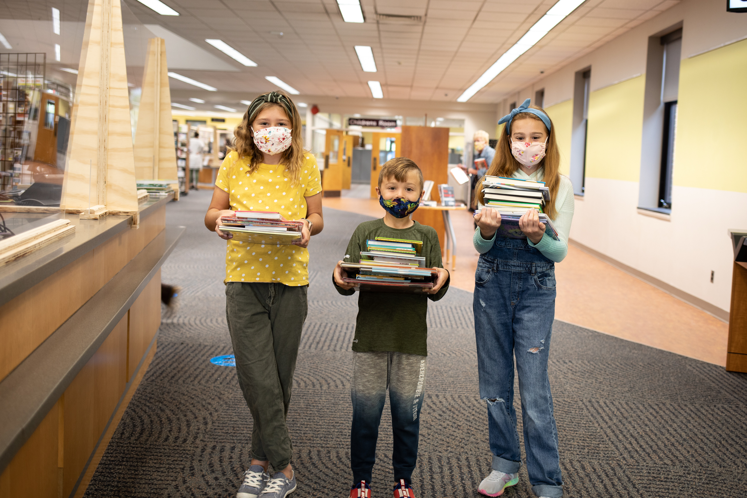 Three masked children hold stacks of books in a library.