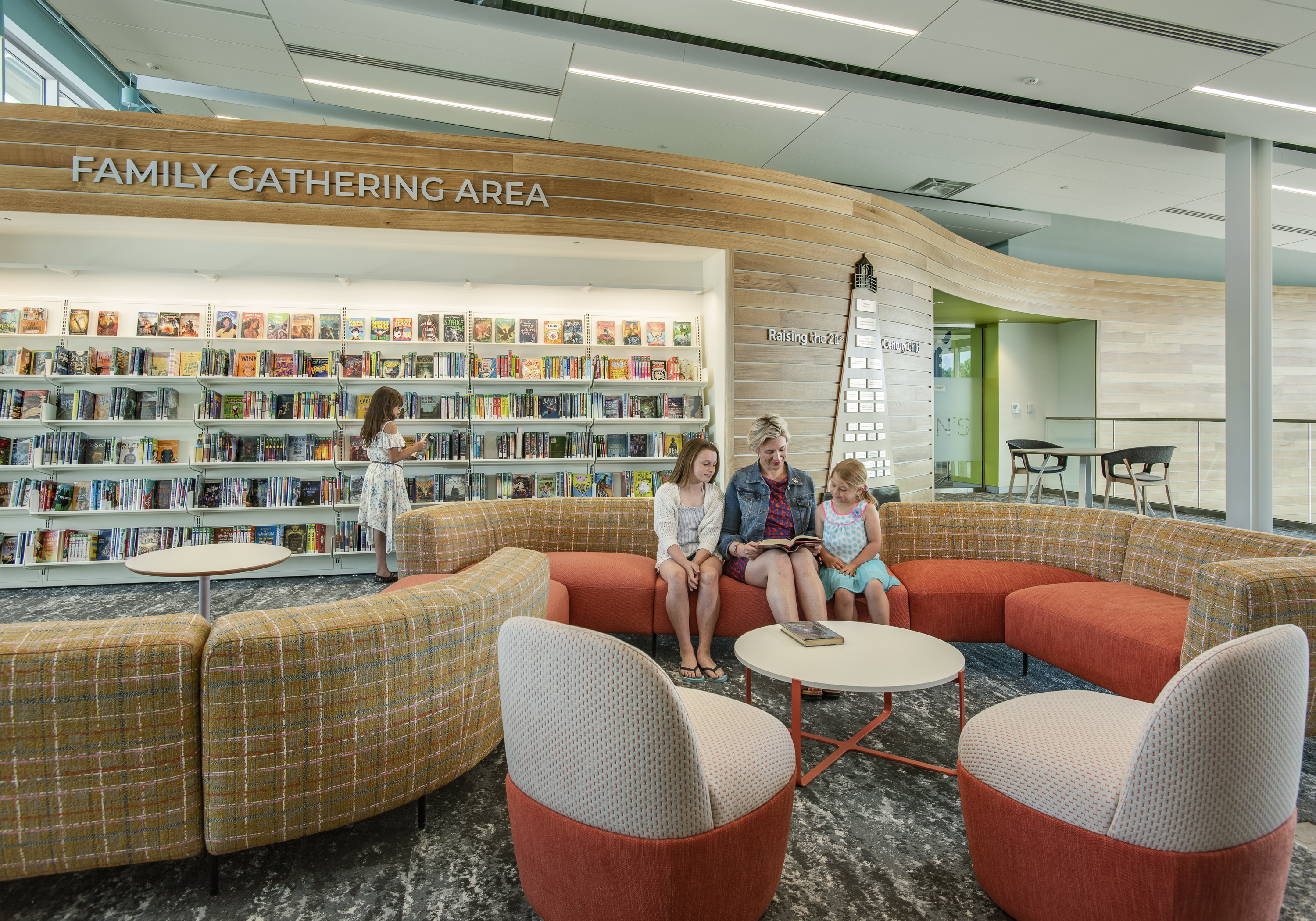 A modern library setting where children can browse for books or sit being read to by an adult on comfortable furniture.
